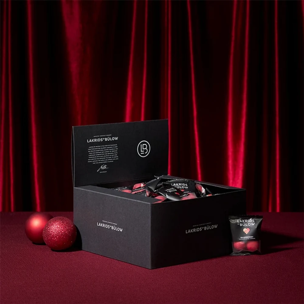 Lakrids by Bülow Box of Minis Crispy Raspberry vor rotem Samtvorhang mit Christbaumkugeln – Firmengeschenk