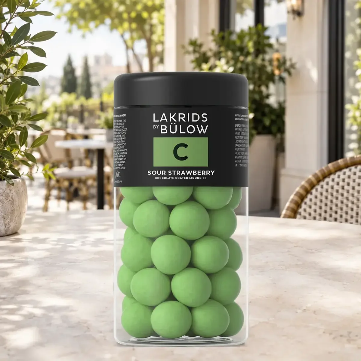 Schoko-Lakritz C Sour Strawberry Lakrids by Bülow, 270 g LAKRIDS BY BÜLOW Sour Strawberry 270 g im schwarzen Lakritzglas als farbintensives Werbegeschenk auf einer hellen Caféterrasse.