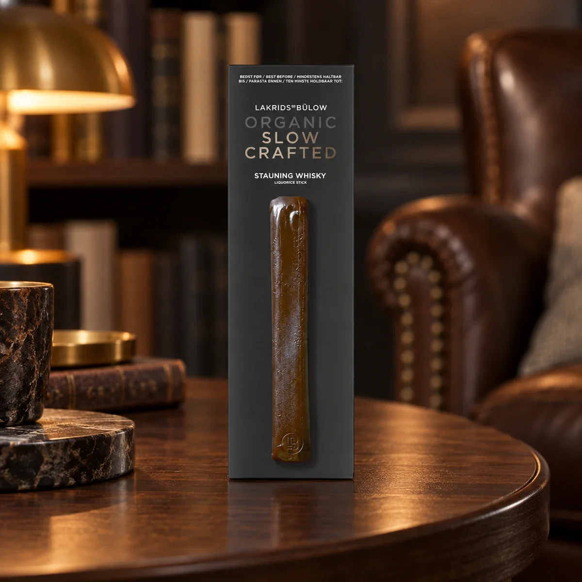 Bio-Lakritz-Stick Stauning Whisky Lakrids by Bülow, 30 g LAKRIDS BY BÜLOW Stauning Whisky Liquorice Stick als exklusives Lakritz-Werbegeschenk auf einem Beistelltisch in einer Bibliothekslounge.