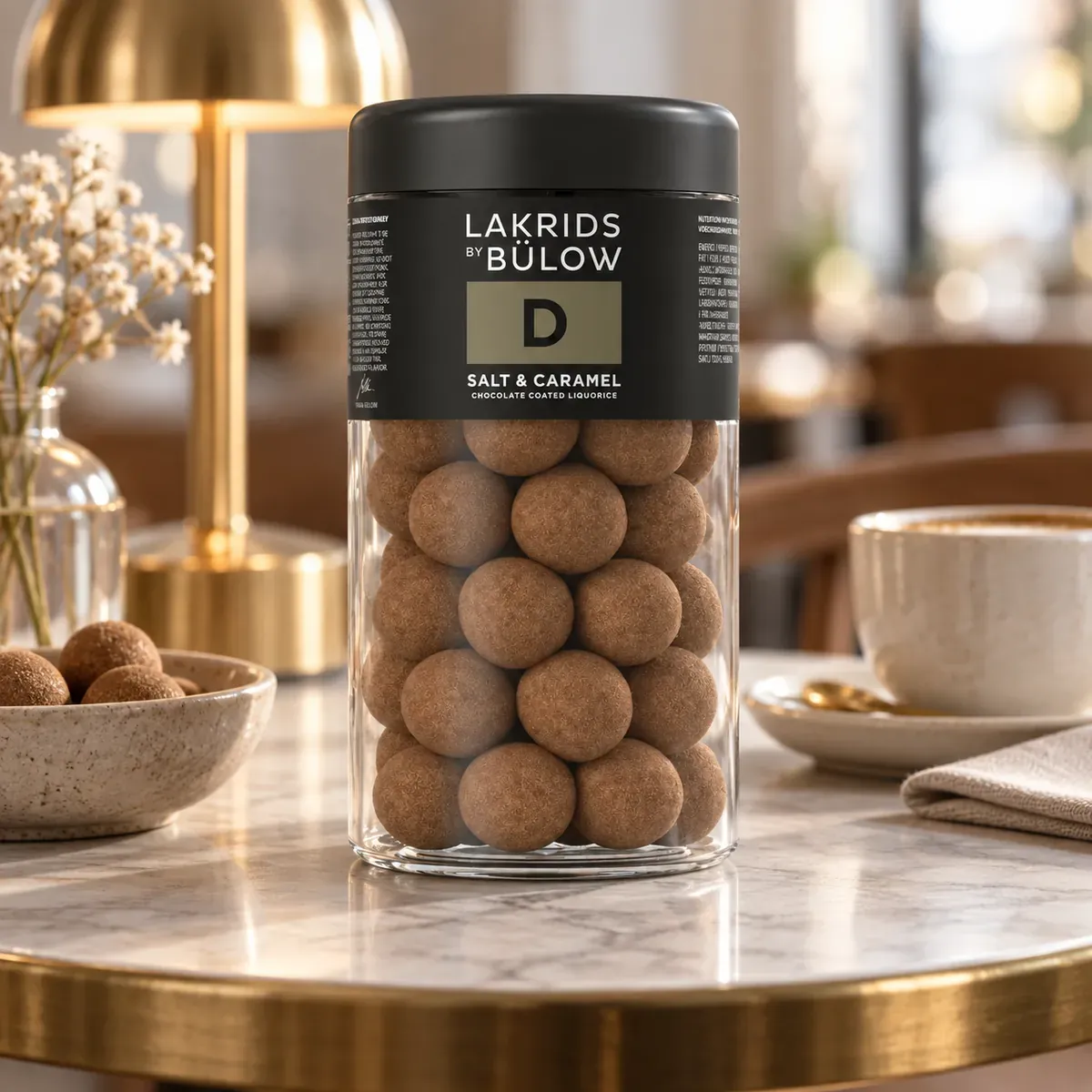 Schoko-Lakritz D Salt & Caramel Lakrids by Bülow, 270 g LAKRIDS BY BÜLOW Salt & Caramel 270 g im schwarzen Glas als edles Lakritz-Werbegeschenk auf einem Café-Beistelltisch.