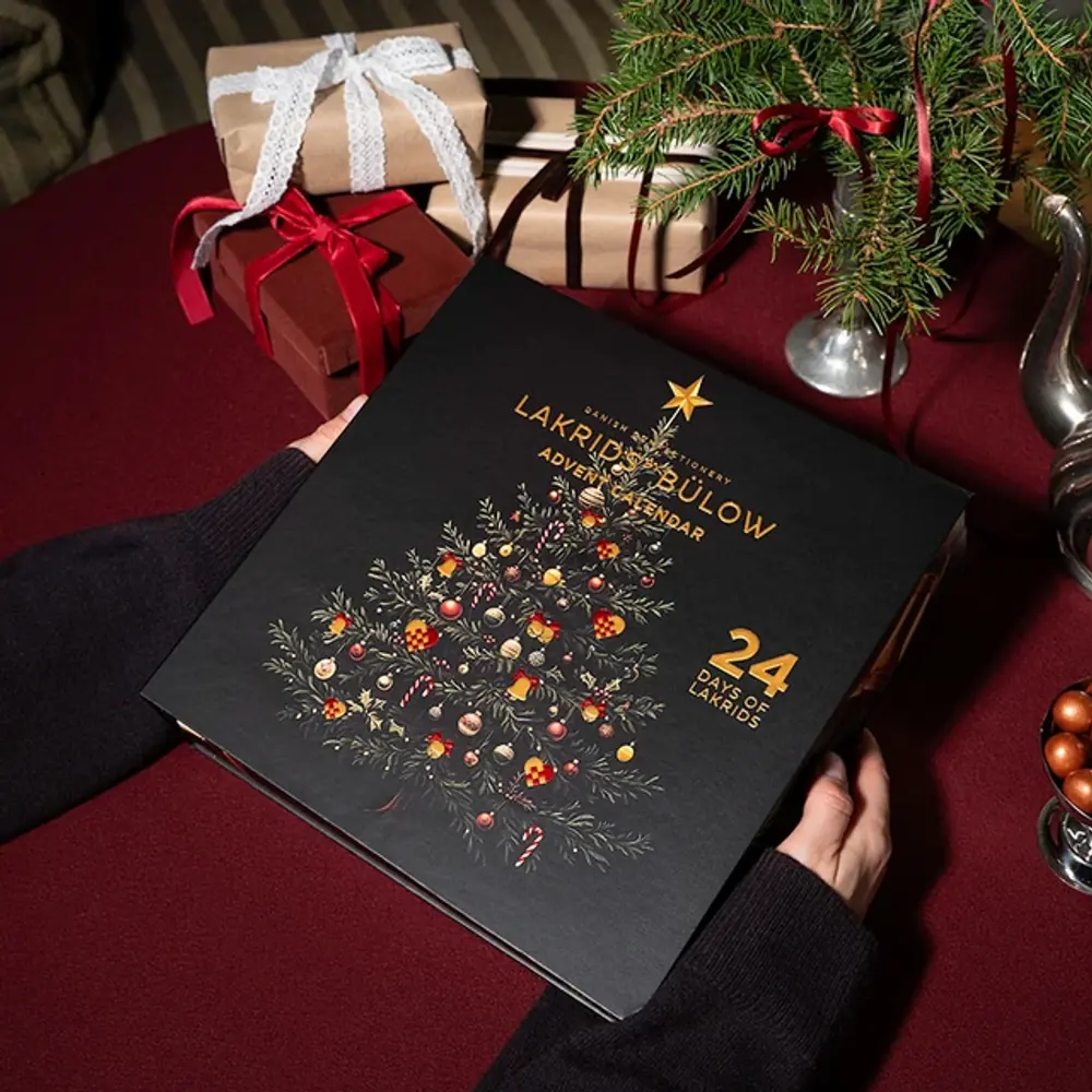 Lakrids by Bülow Adventskalender in Händen vor Geschenken auf rotem Tisch – Adventsgeschenk für Kunden